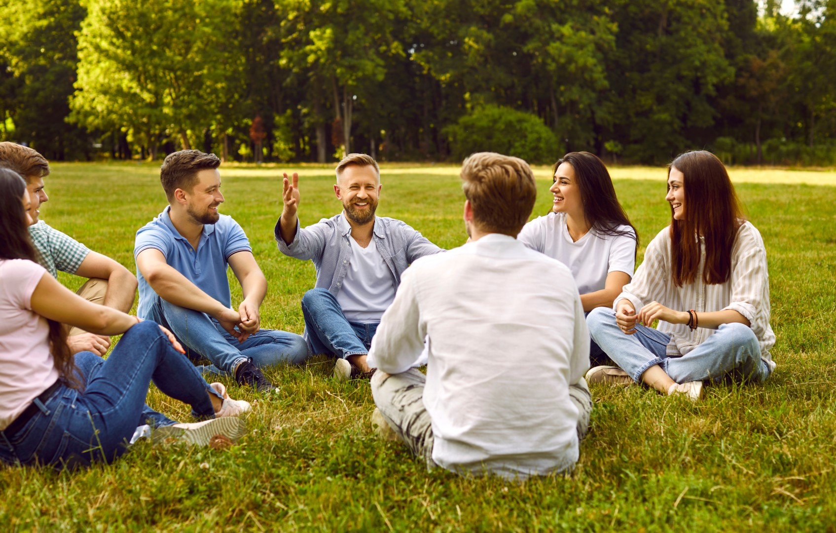 Group of young people communicate sitting on grass in park on summer sunny day.